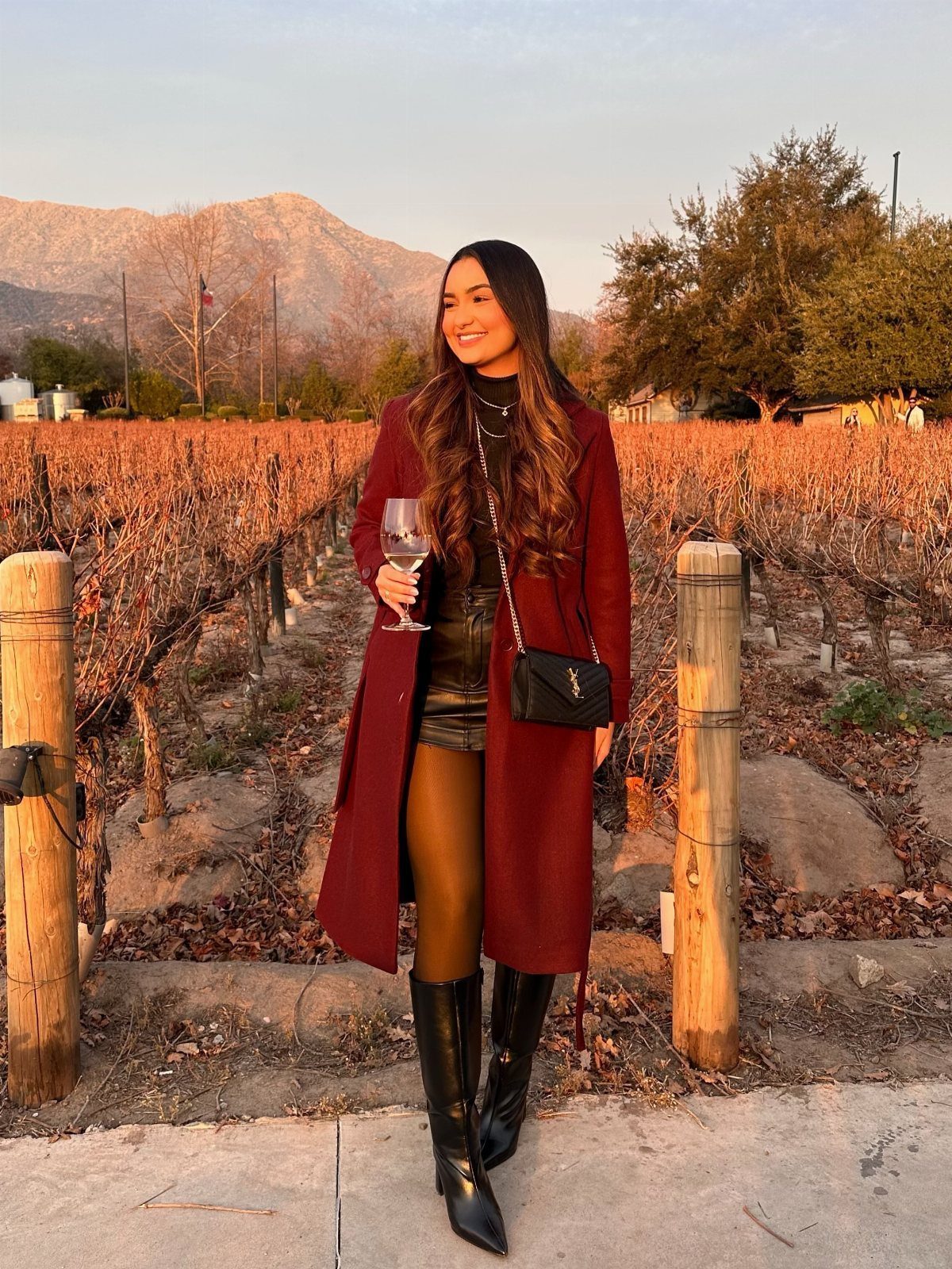 Woman in a burgundy coat and black boots standing in a vineyard during sunset.