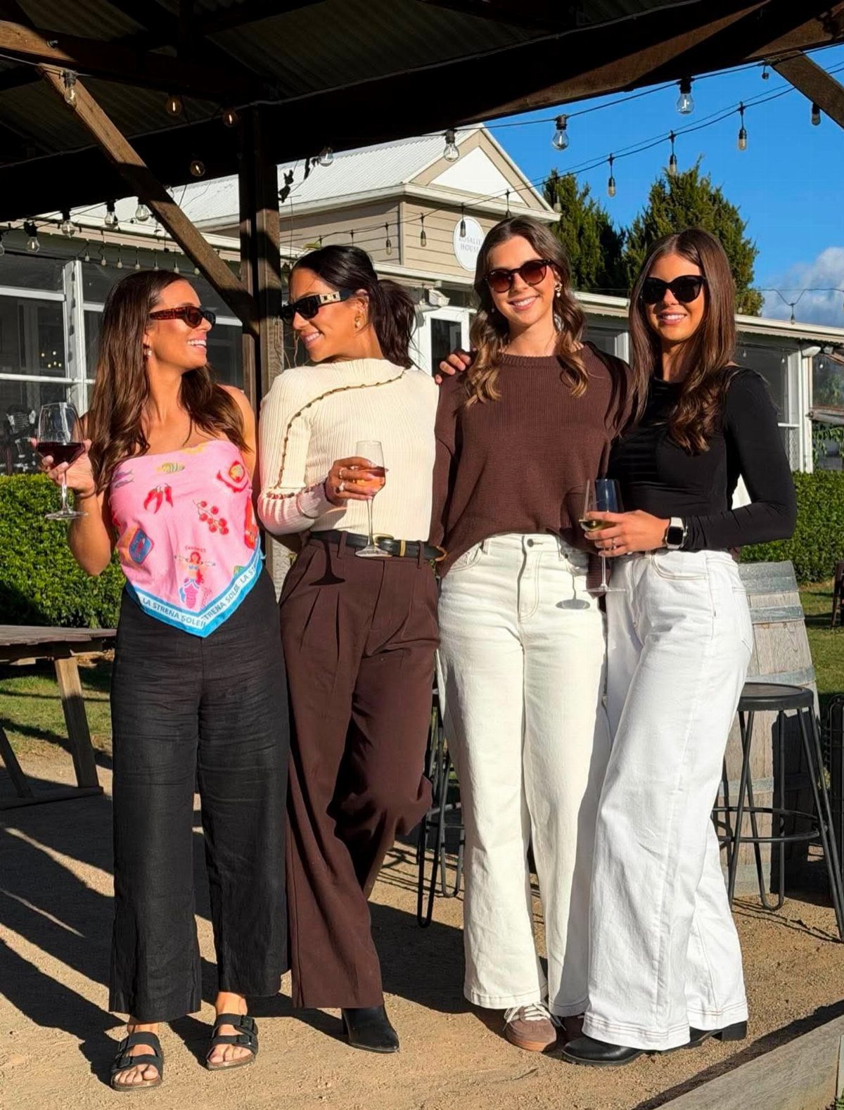 Four women smiling and holding wine glasses while wearing stylish, casual outfits outdoors.