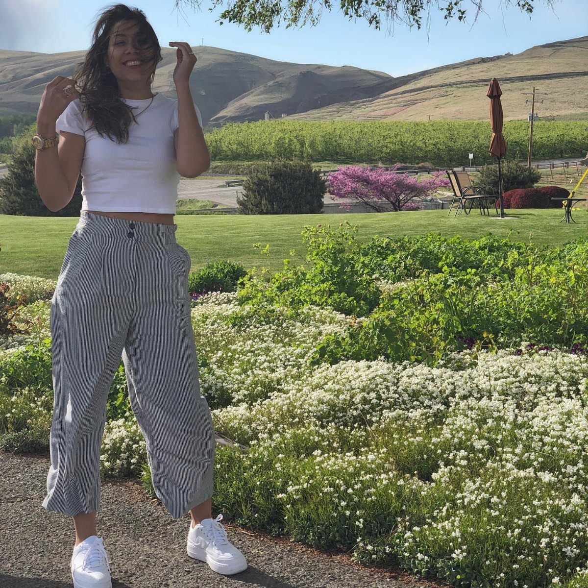 A woman in striped trousers and white sneakers posing in a sunny vineyard landscape.