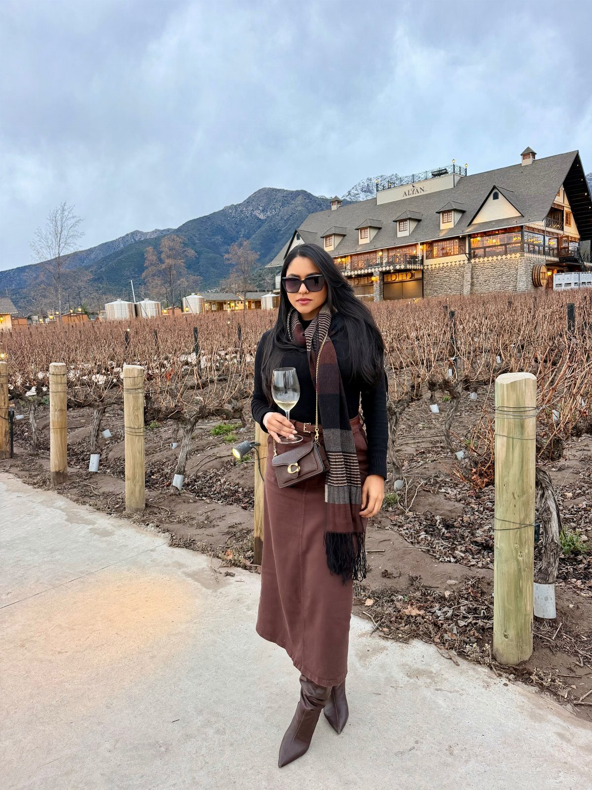 Woman in a vineyard wearing a scarf, long sleeves, and boots under an overcast sky.