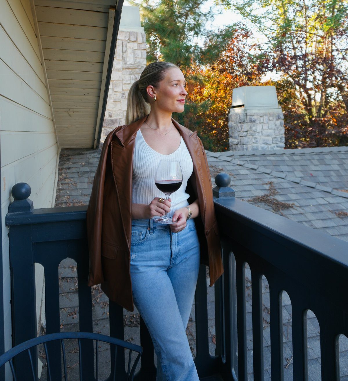 A woman in a white tank top and blue jeans wearing a brown leather jacket while holding wine.