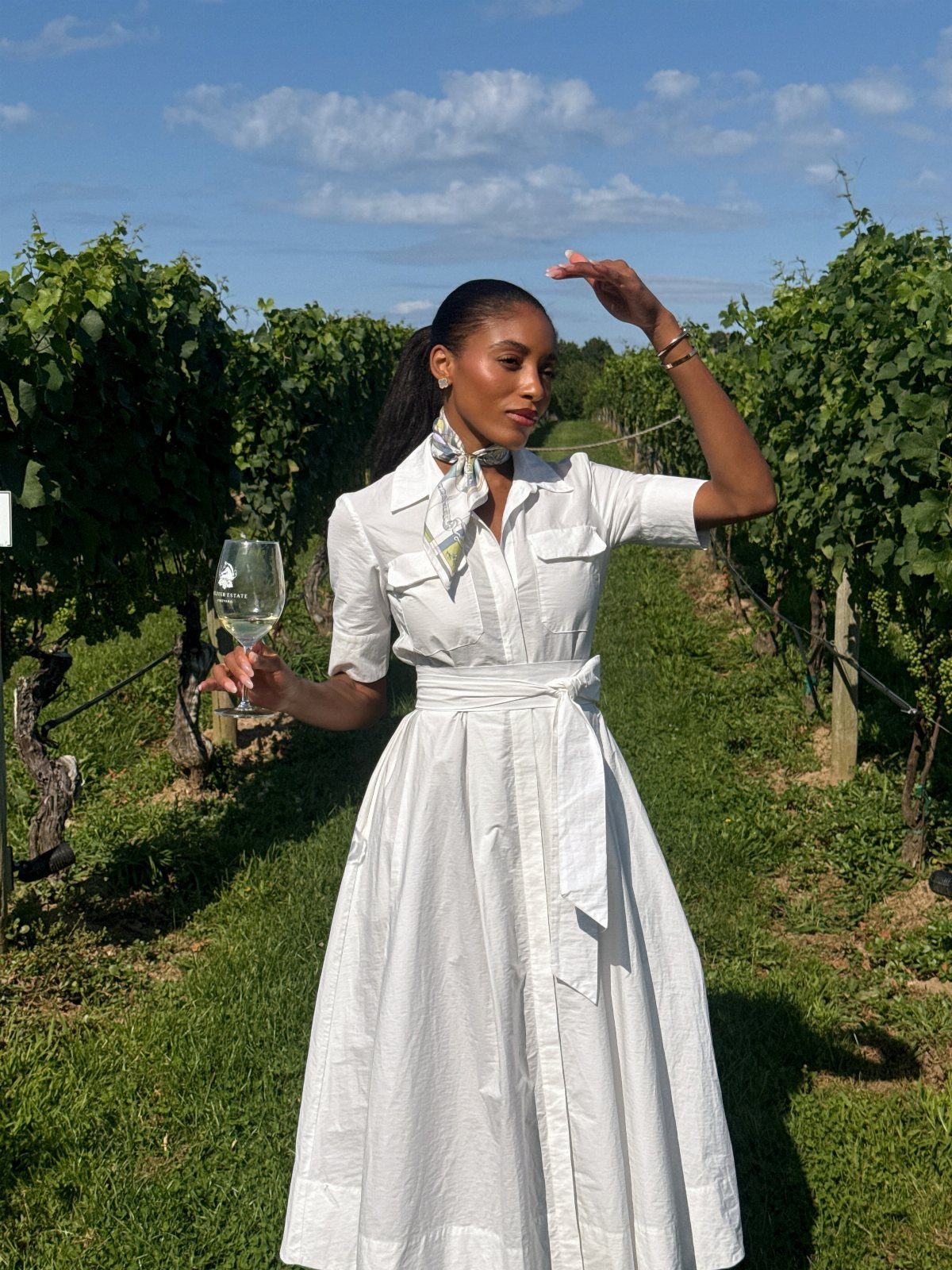Woman in a white midi dress and neck scarf standing in a sunny vineyard holding wine.