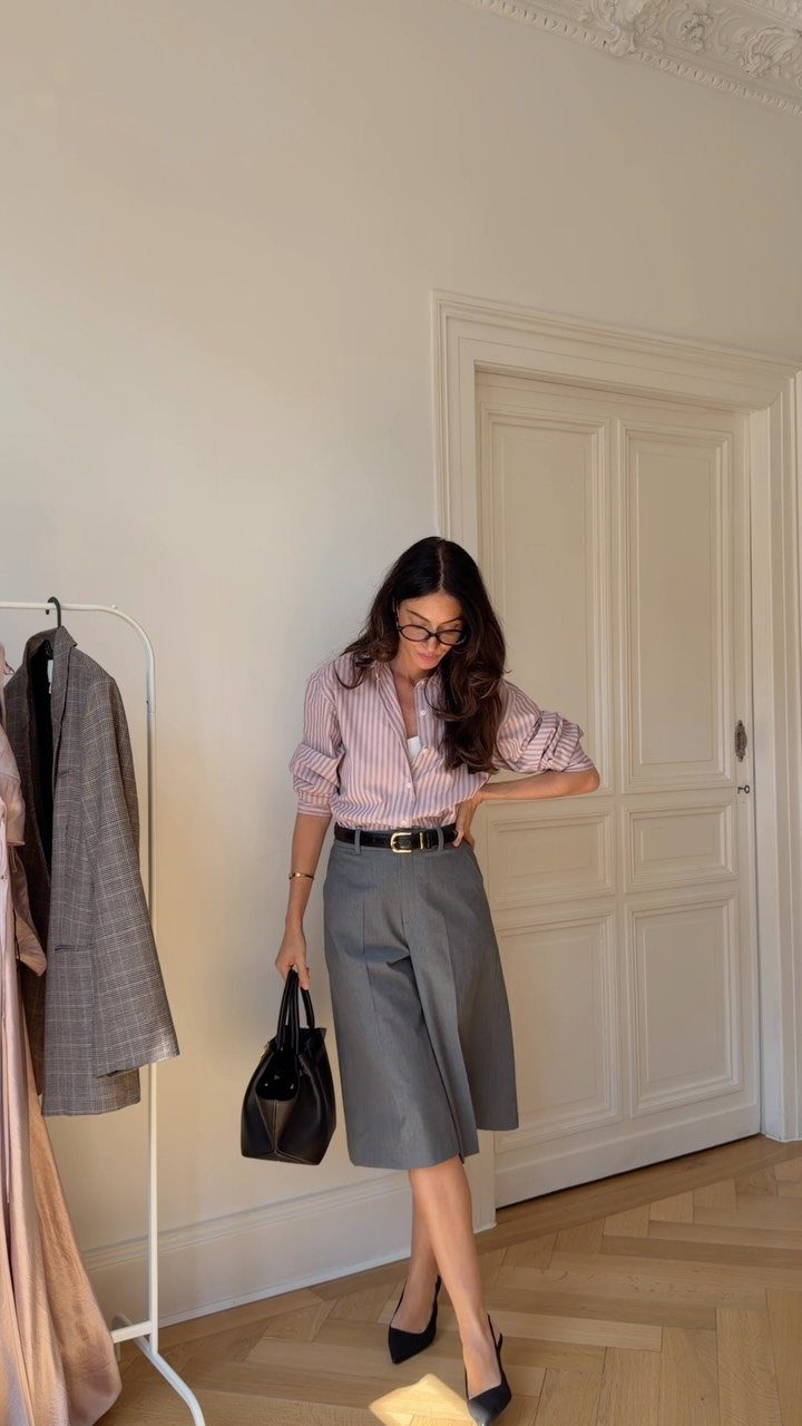Woman wearing a pink striped shirt, black belt, and grey culottes standing in a room.