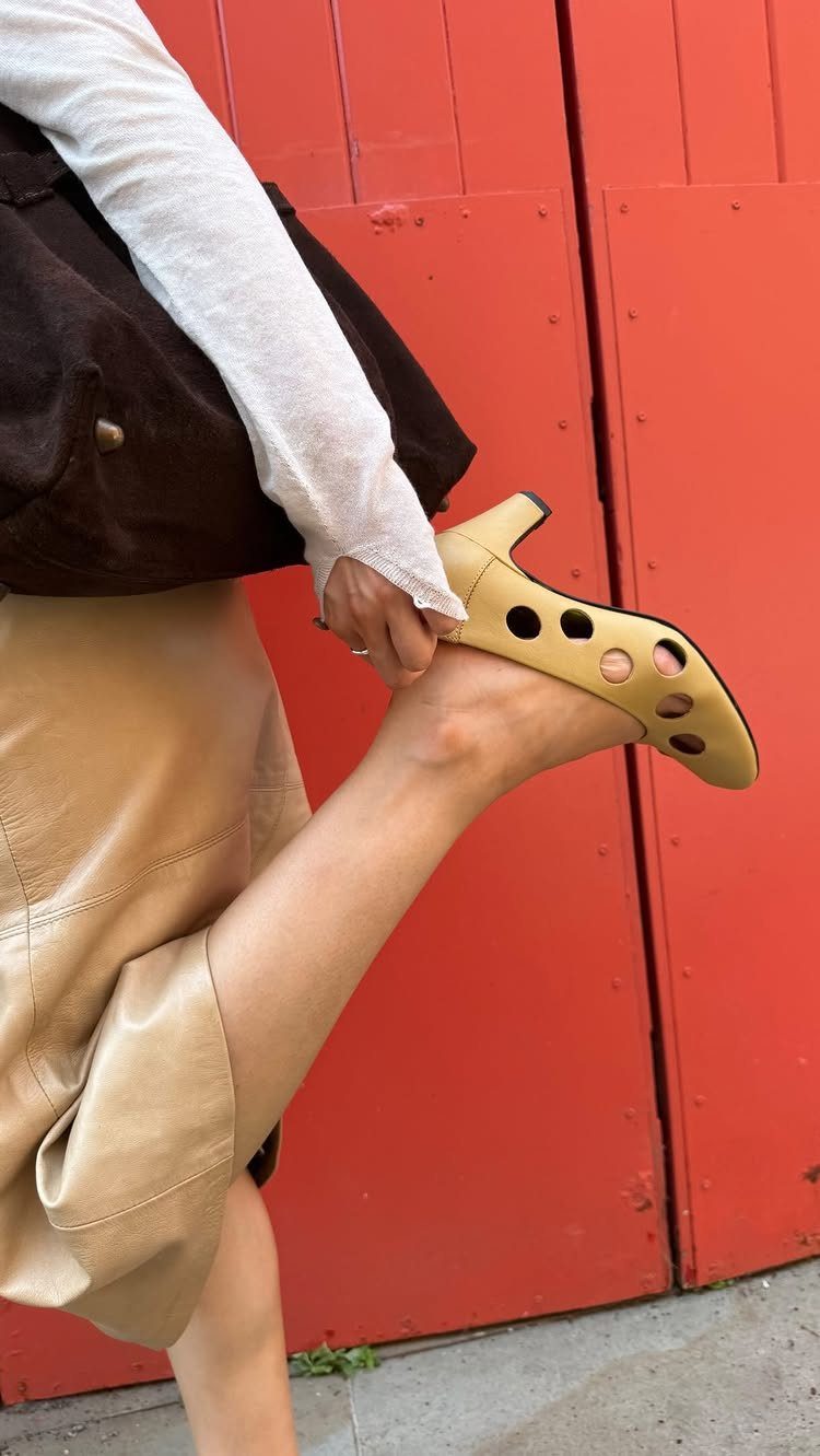 A person holding a beige perforated leather pump shoe against a red wooden door.