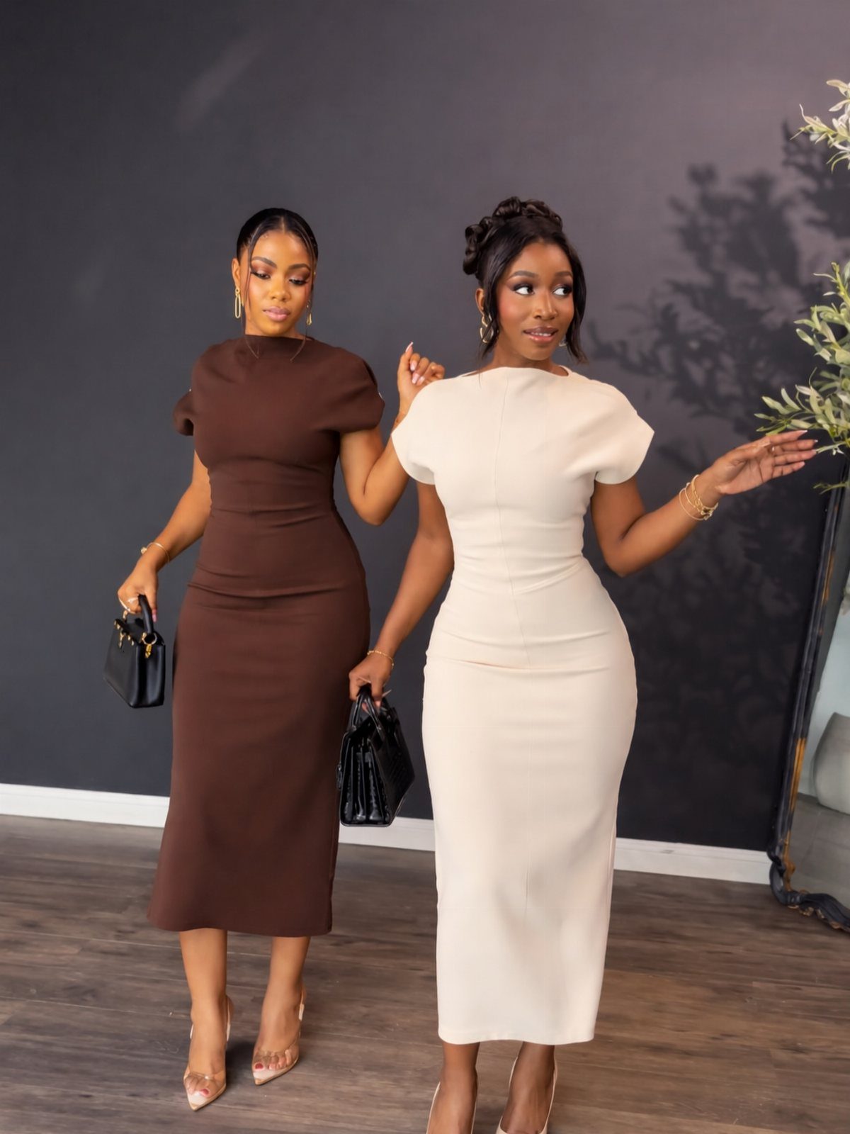 Two women posing in elegant, short-sleeved midi dresses and high heels against a dark backdrop.