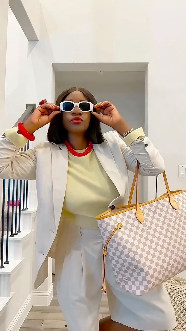 Woman in a white blazer, yellow top, and white trousers holding sunglasses and a large tote bag.