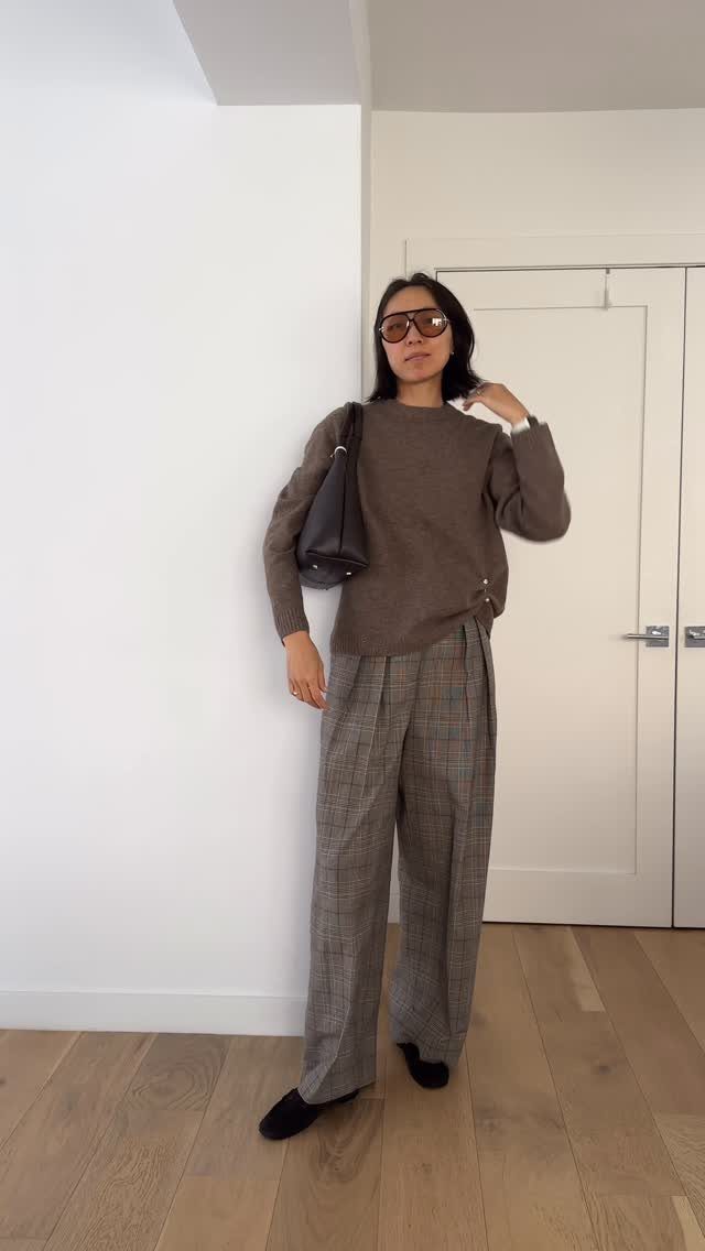 Woman posing in a brown sweater, plaid trousers, and sunglasses against a white wall.