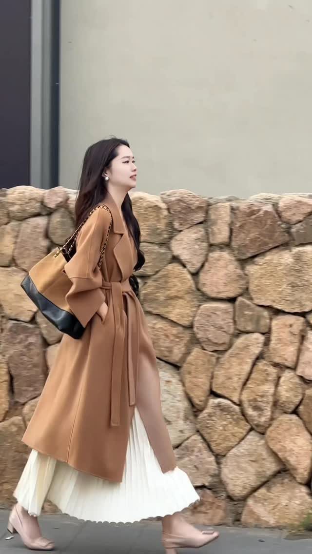 Woman walking in a camel coat, white pleated skirt, and beige heels near a stone wall.