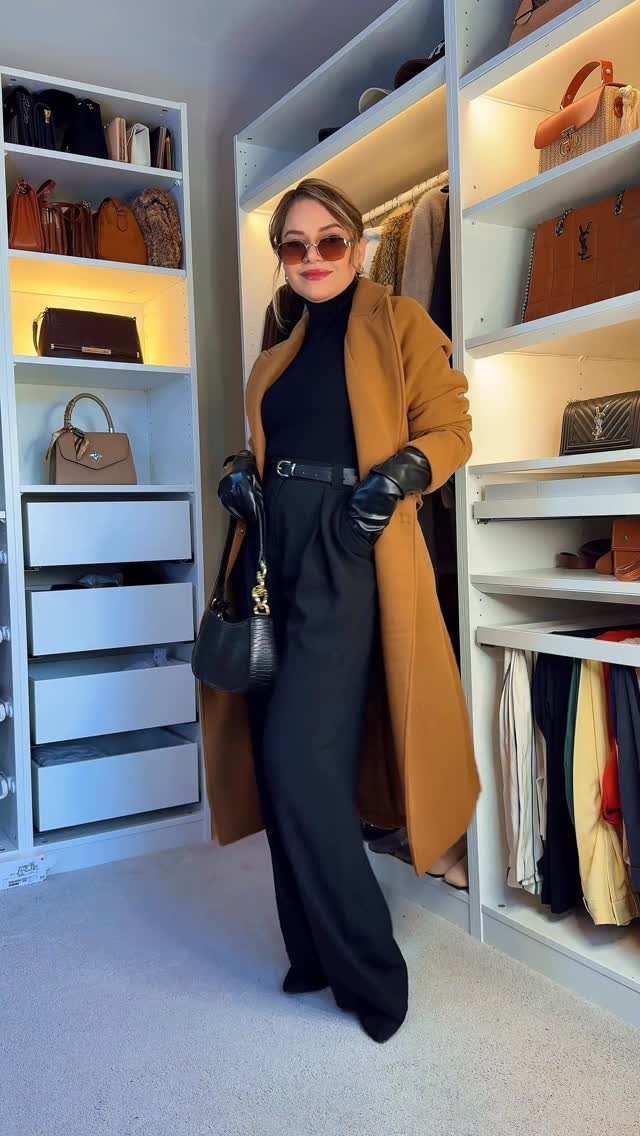 Woman wearing a long camel coat, black turtleneck, and black trousers standing in a closet.
