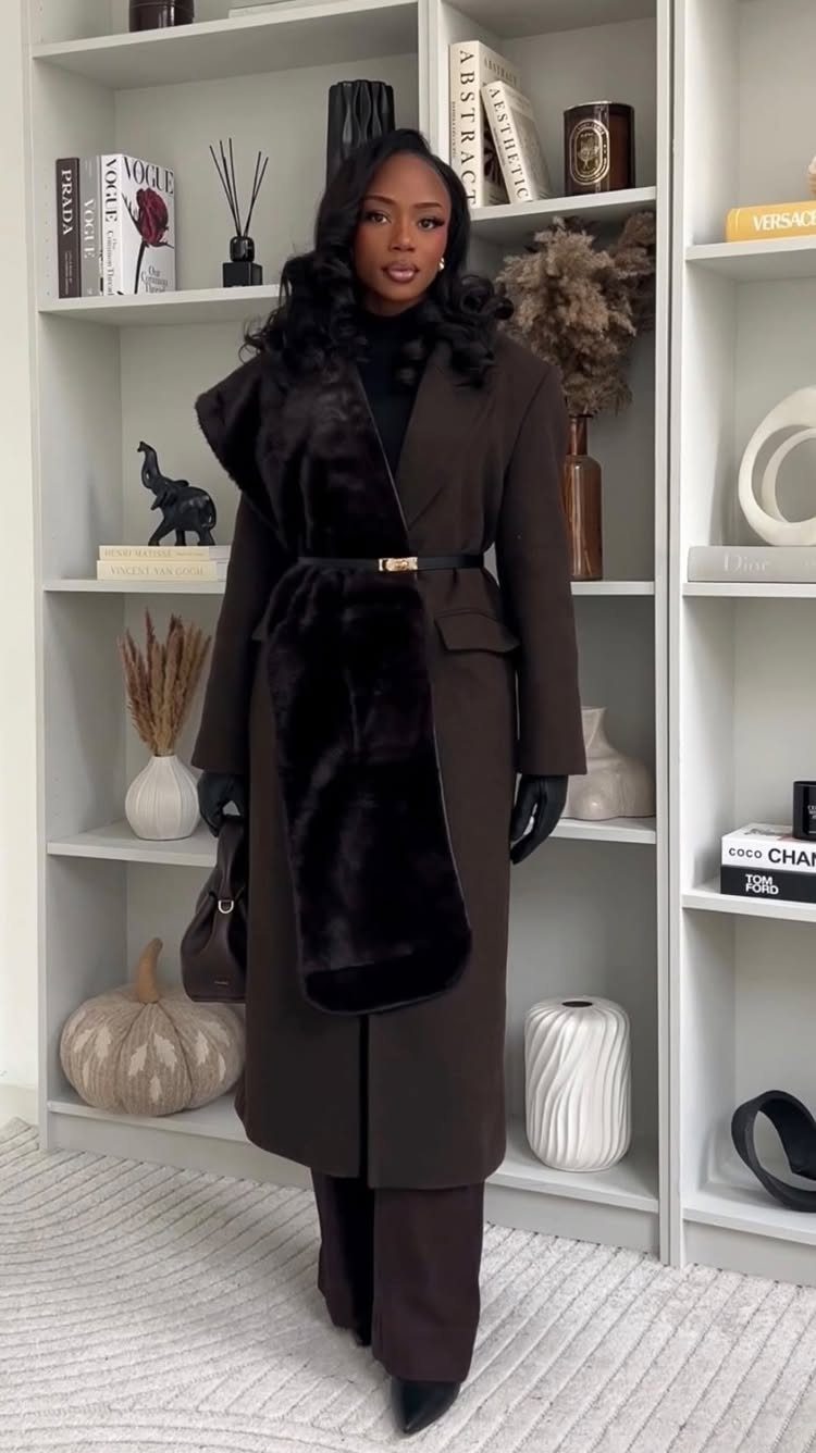 A woman wearing a long brown coat with a black fur stole and dark trousers.