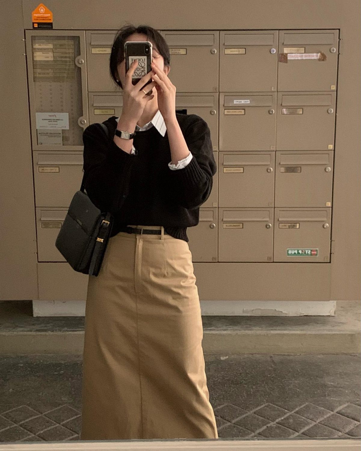 Woman in black sweater and tan skirt taking a mirror selfie with a black bag.