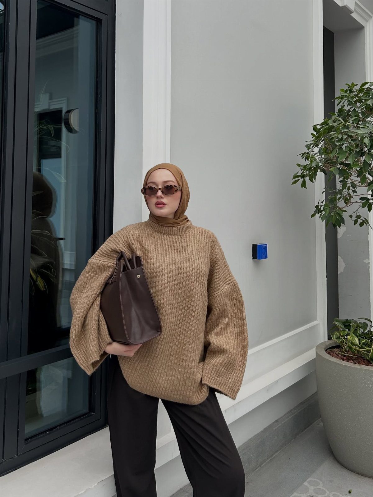 Woman in a camel knit sweater and black trousers holding a brown leather tote bag.