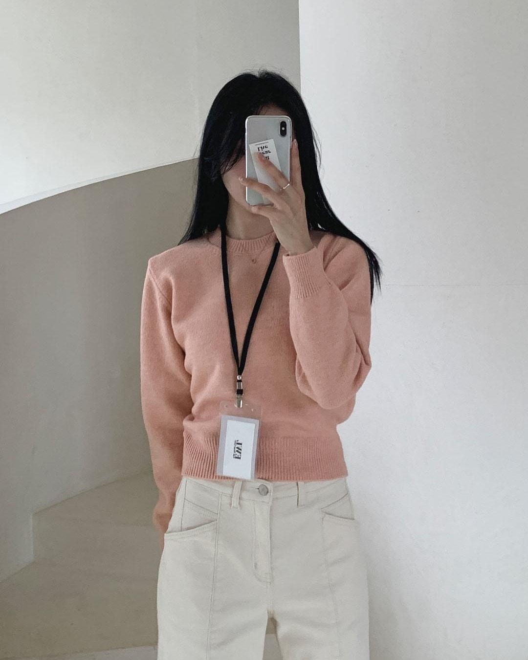 Woman in a peach sweater and white trousers posing for a mirror selfie.