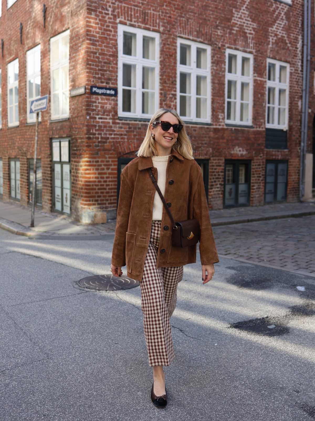 Smiling woman in a brown suede jacket and checkered trousers walking on a city street.
