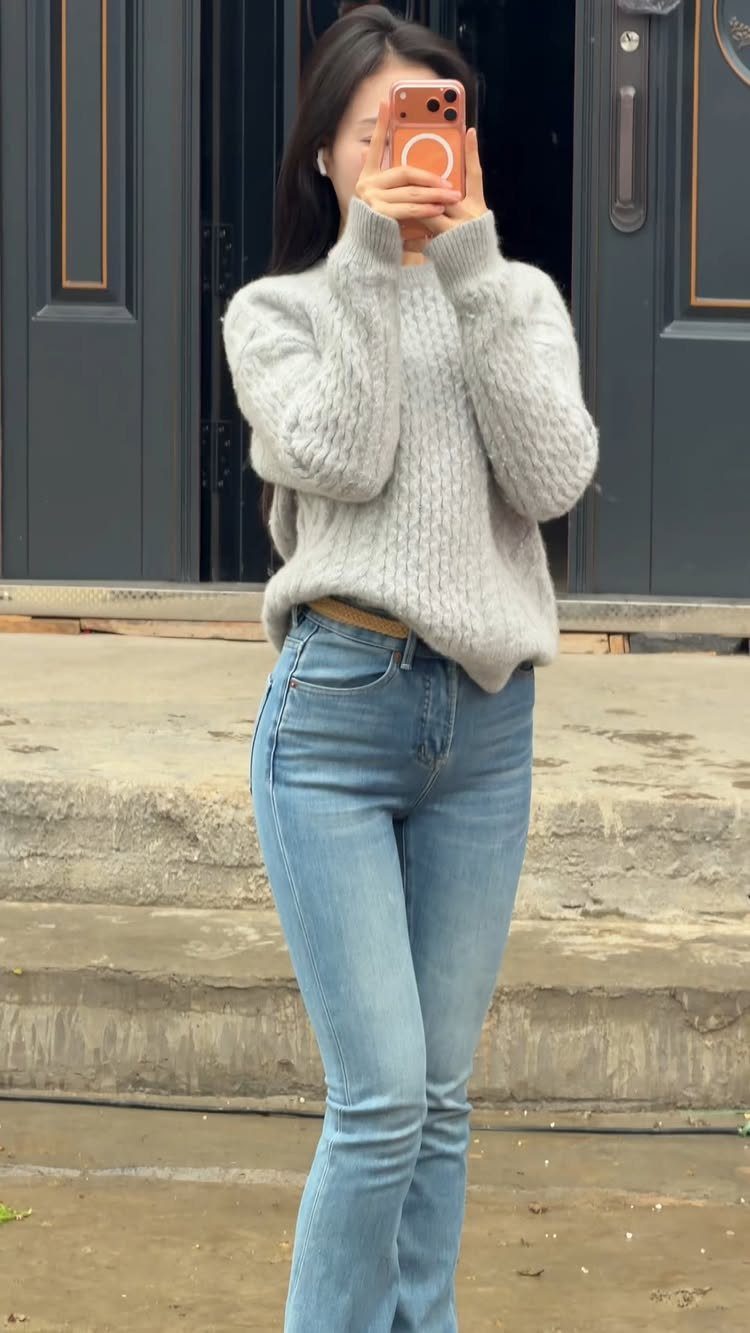 Woman wearing a grey cable-knit sweater and high-waisted blue jeans standing outdoors.
