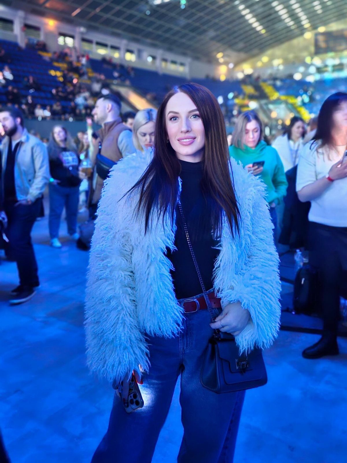 A woman wearing a black turtleneck, blue jeans, and a large white faux fur coat in an arena.