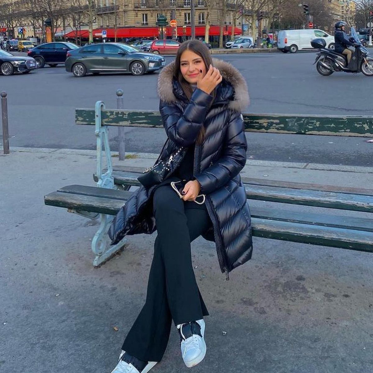 Woman in a long black puffer coat, black flared pants, and white sneakers sitting on a bench.