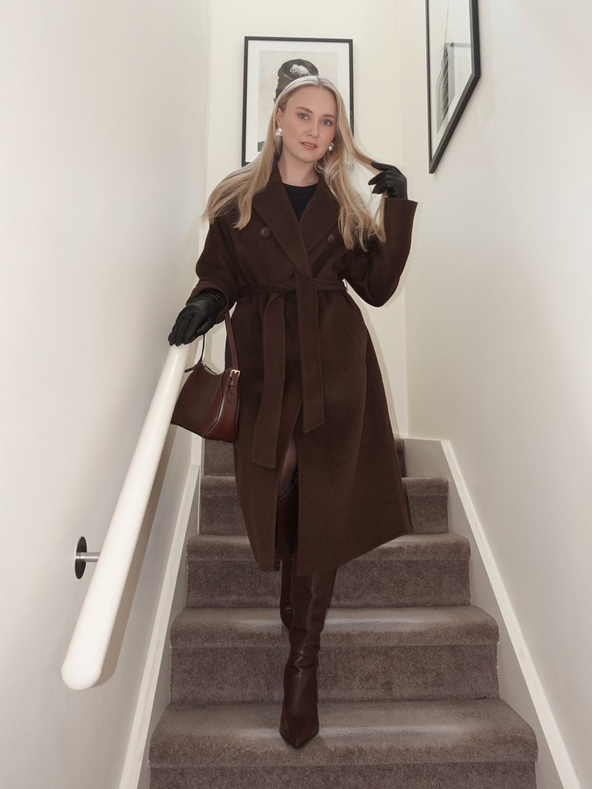 Blonde woman in a long brown belted coat and tall boots posing on a staircase.