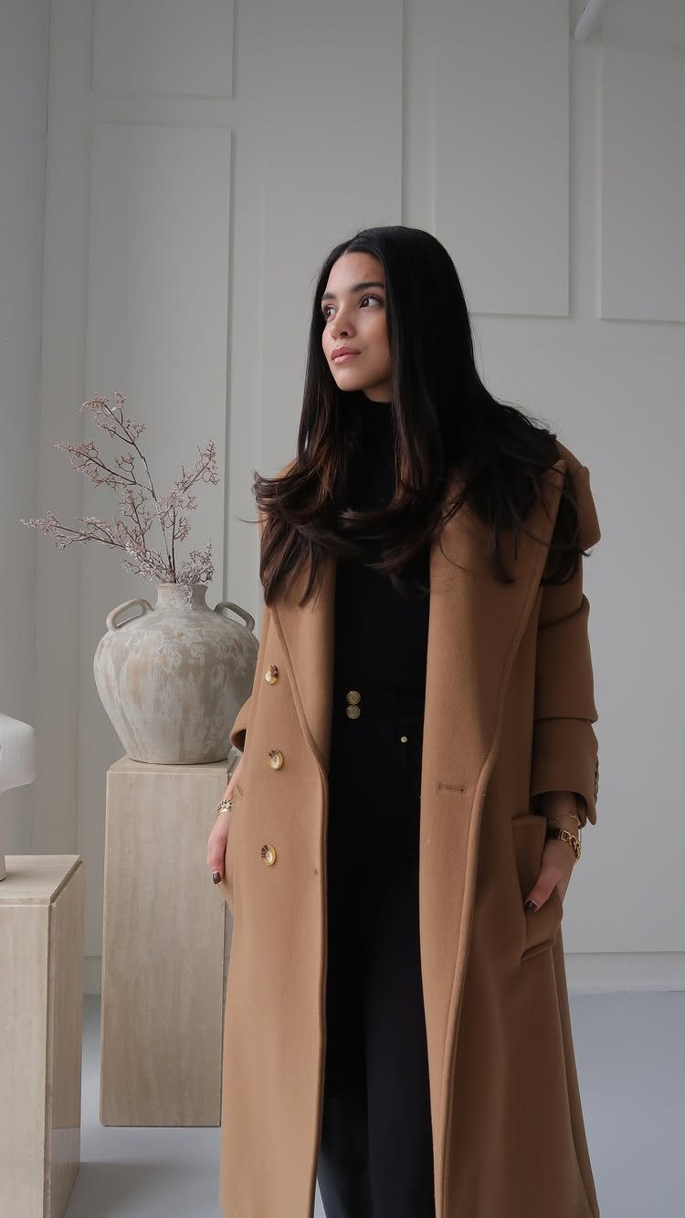 Woman wearing a long camel coat and black turtleneck standing in a minimalist white room.