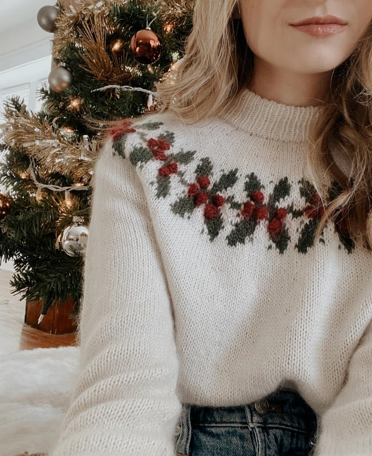 Woman wearing a cream knit sweater with holly berry patterns in front of a Christmas tree.
