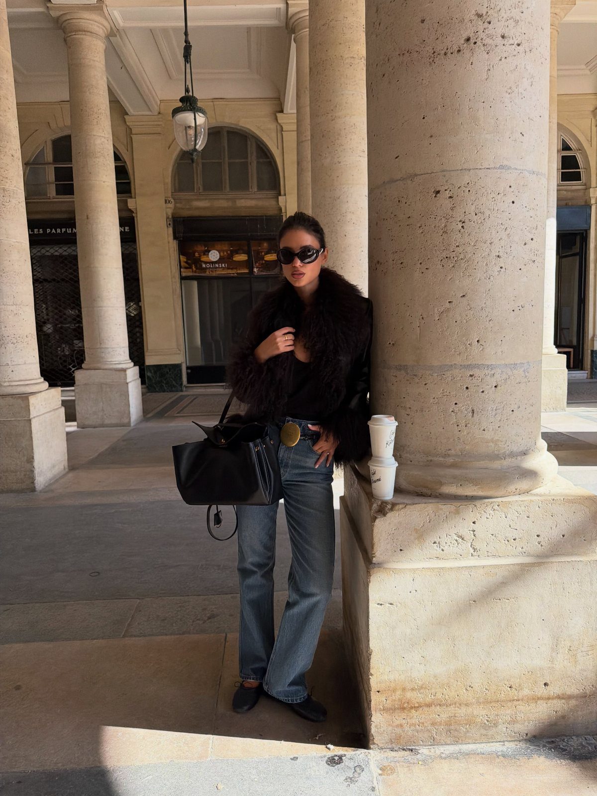 Woman in a black fur coat and blue jeans leaning against a stone column.