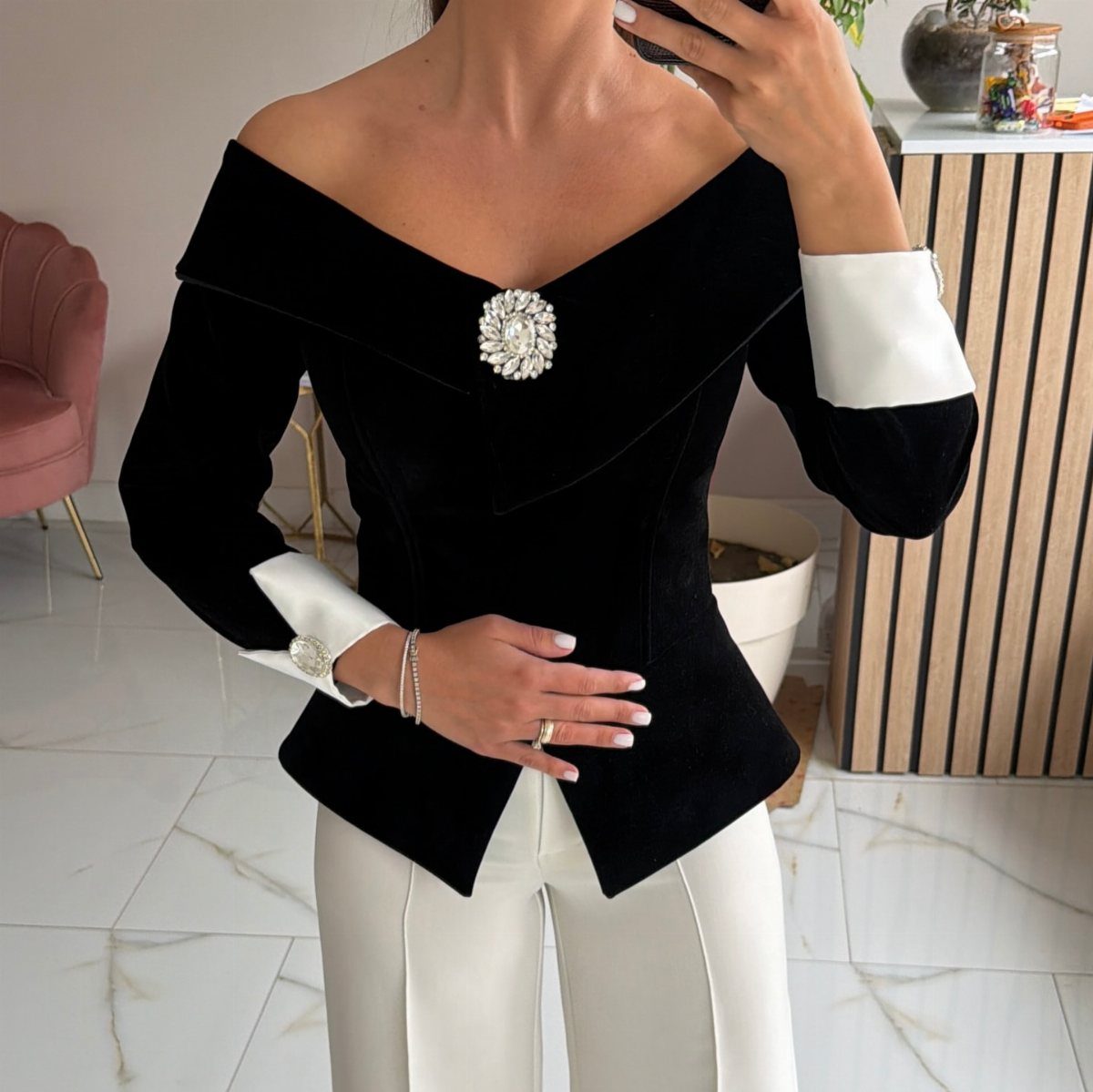 Woman in a black velvet off-the-shoulder top and white trousers taking a mirror selfie.