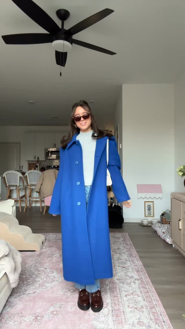 Woman in a long bright blue coat, white turtleneck, and jeans standing in a living room.