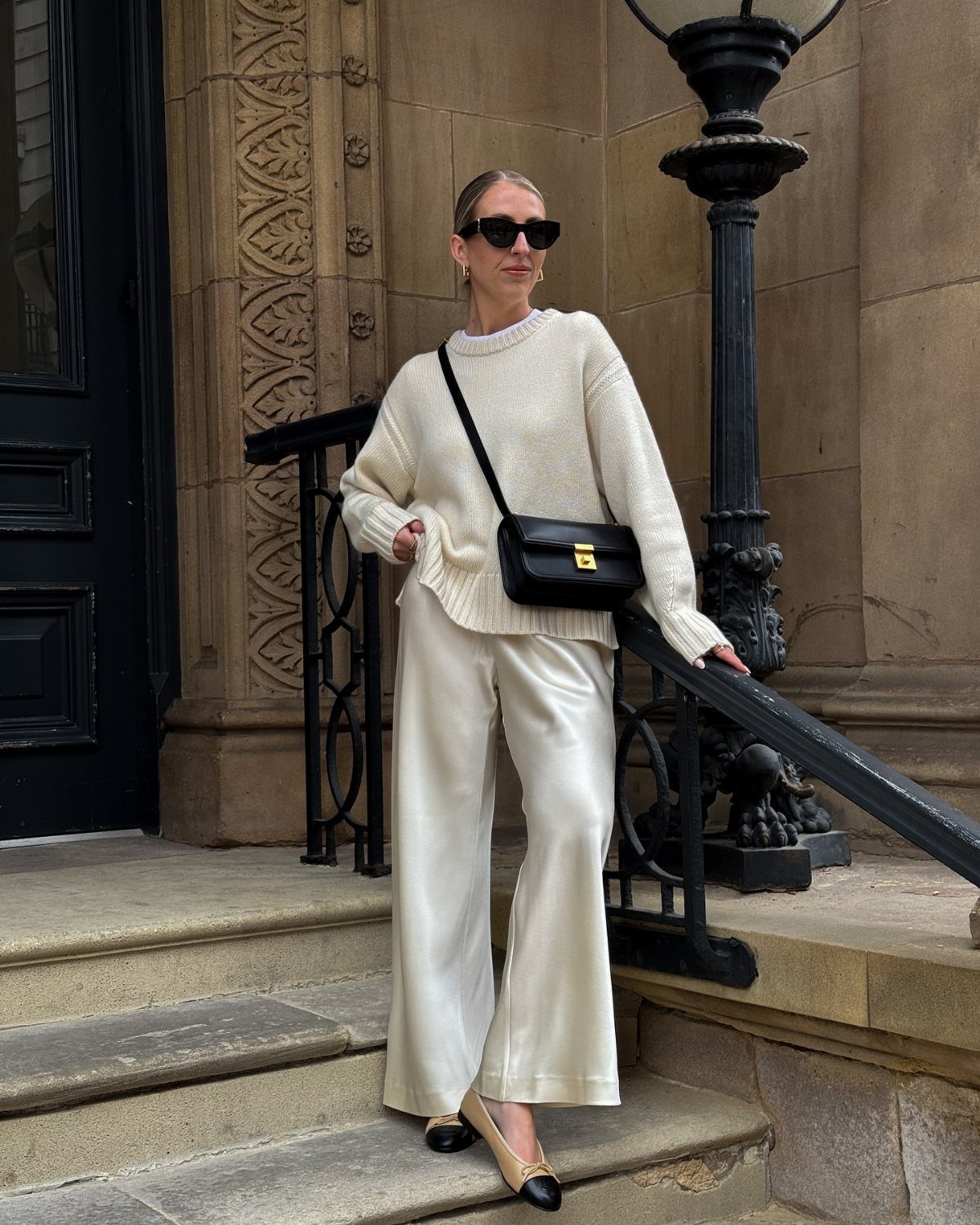 Woman in a cream knit sweater and satin trousers posing on stone steps.