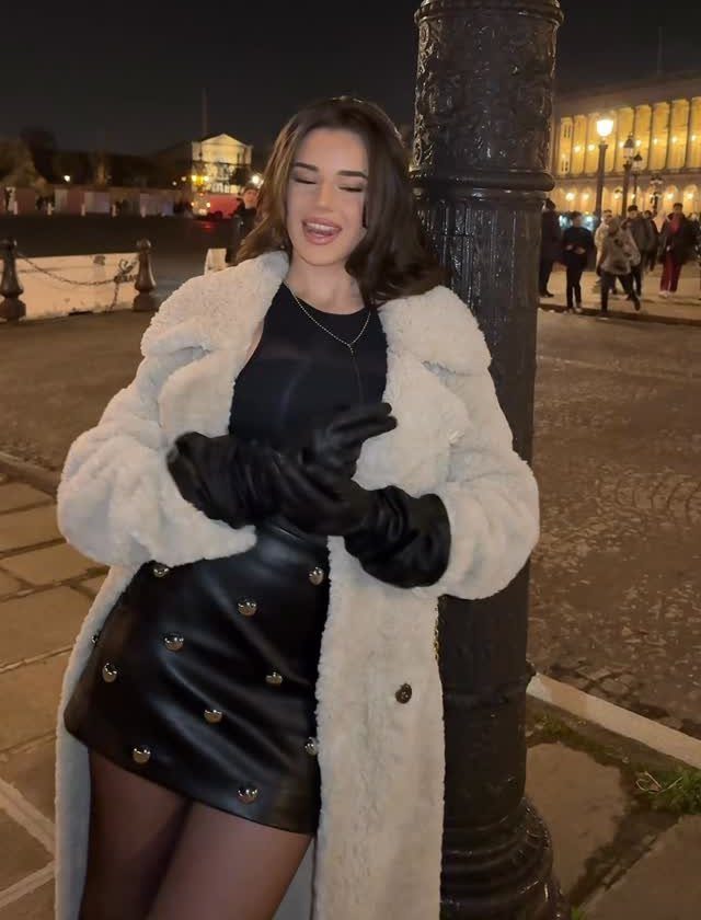 Woman in a black studded mini skirt and white faux fur coat posing at night.