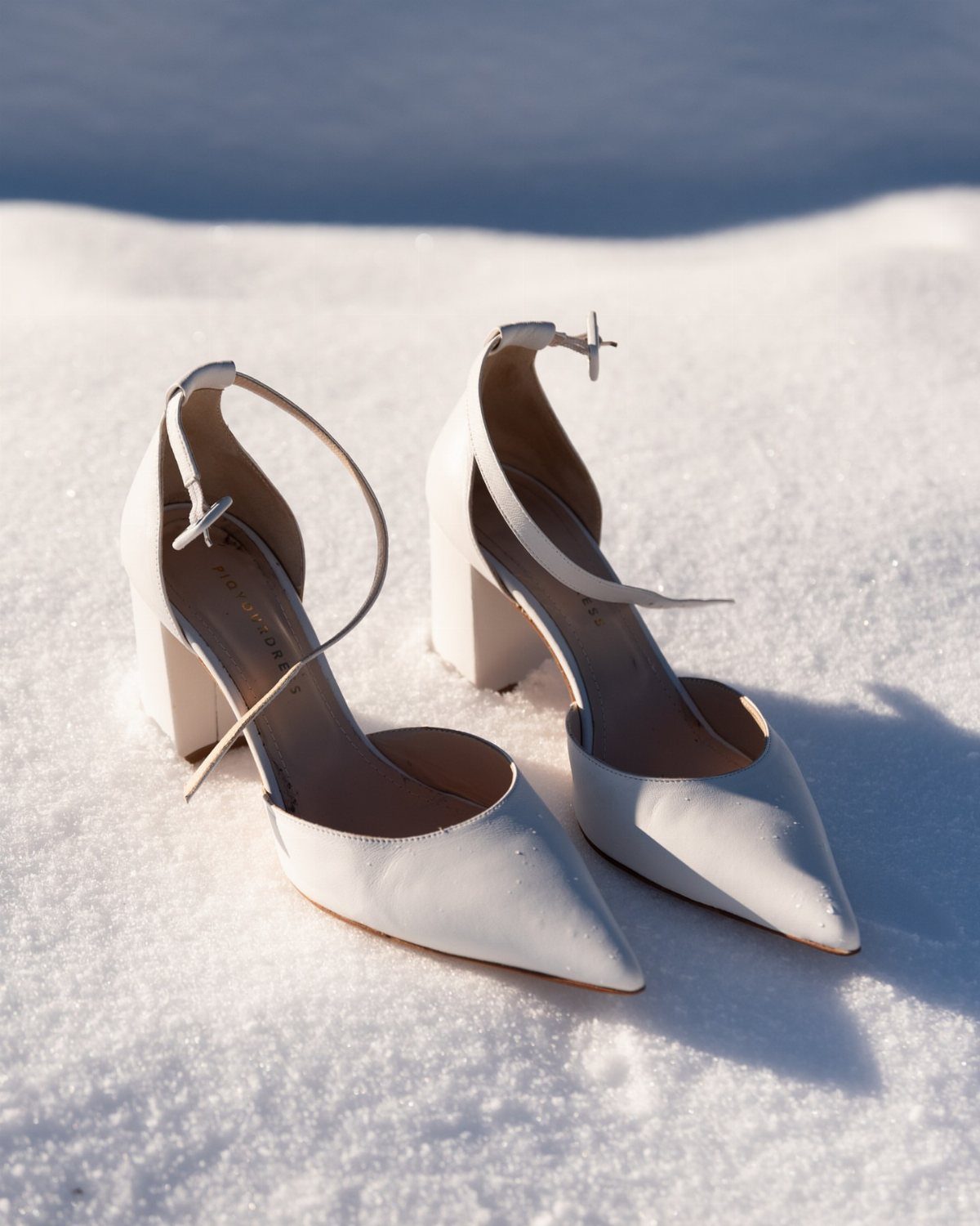 White pointed-toe block heel pumps with ankle straps sitting in white snow.