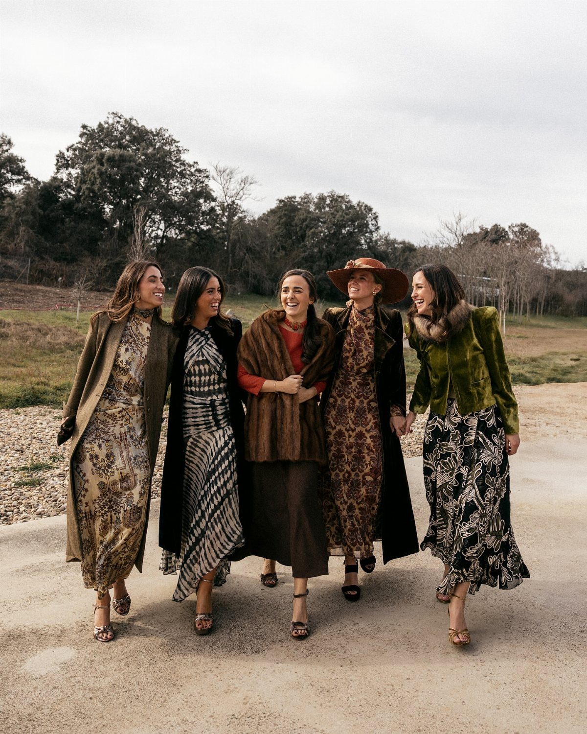 Five women walking outdoors wearing elegant winter wedding guest outfits and long coats.