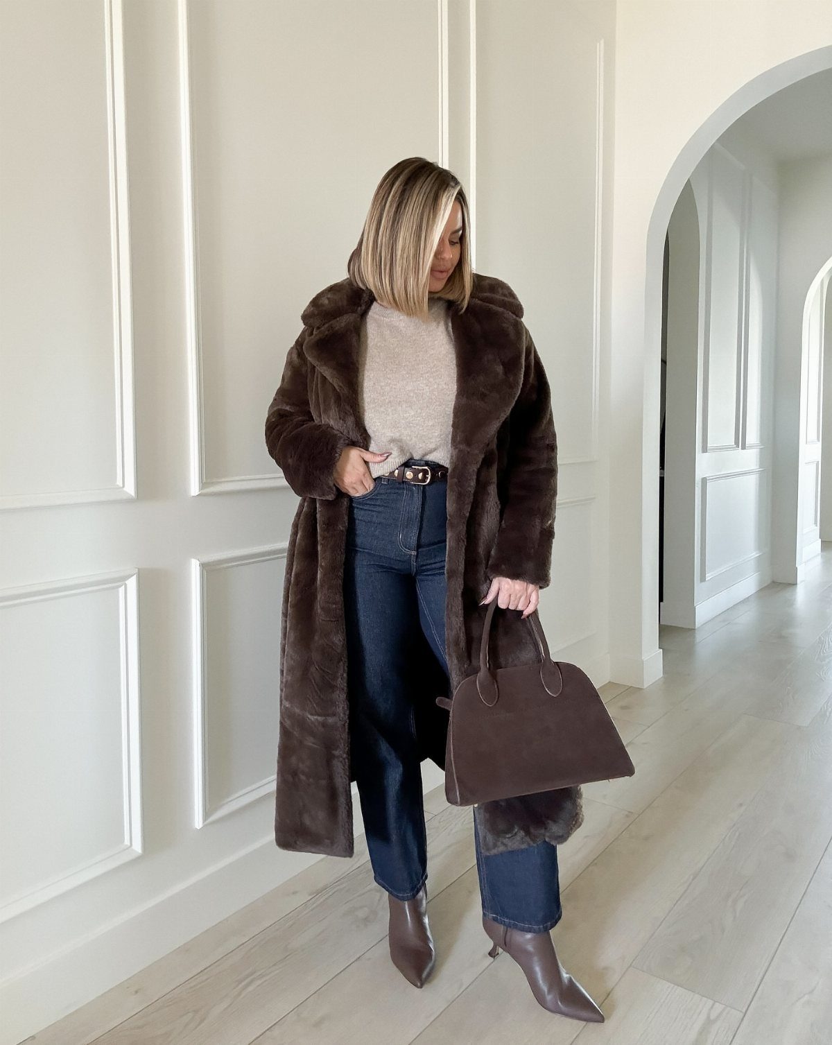 Woman wearing a long brown fur coat, beige sweater, and dark wash jeans walking in a hallway.