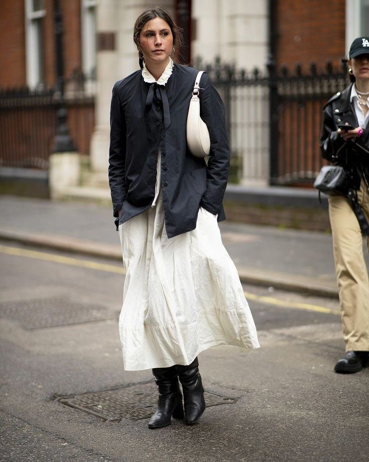 A woman walking in a city wearing a navy jacket, white skirt, and black boots.