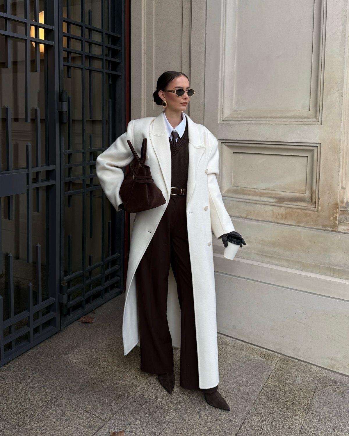 Woman in a long white coat and dark brown trousers standing by a building.