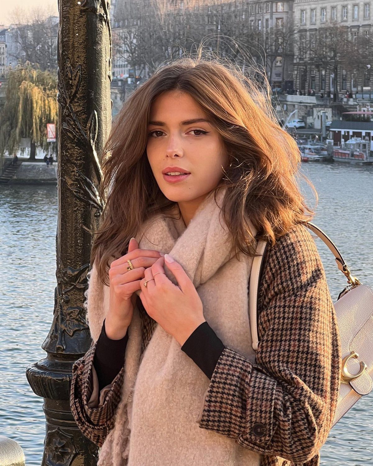 A woman wearing a houndstooth coat and a beige scarf standing by a river.