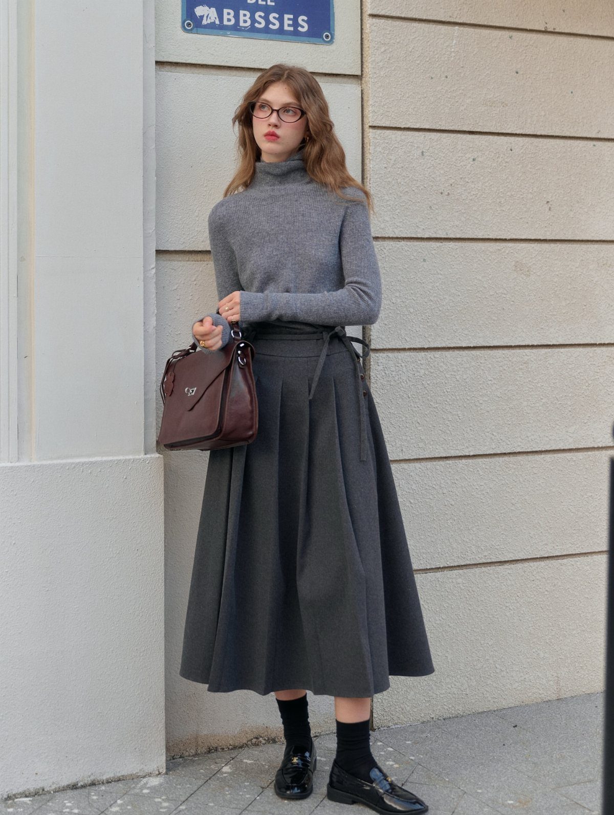 Woman wearing a grey turtleneck sweater and a dark grey pleated midi skirt with black loafers.
