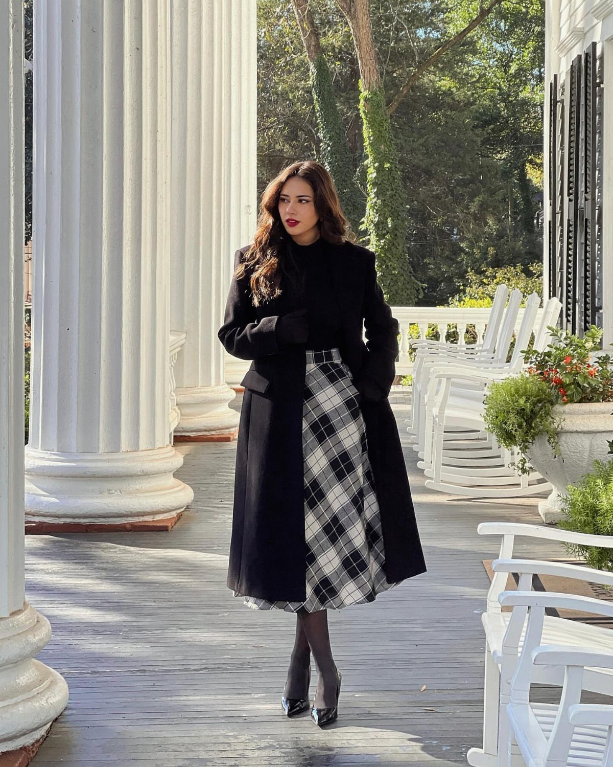 Woman wearing a black coat, black turtleneck, and plaid midi skirt standing on a white porch.