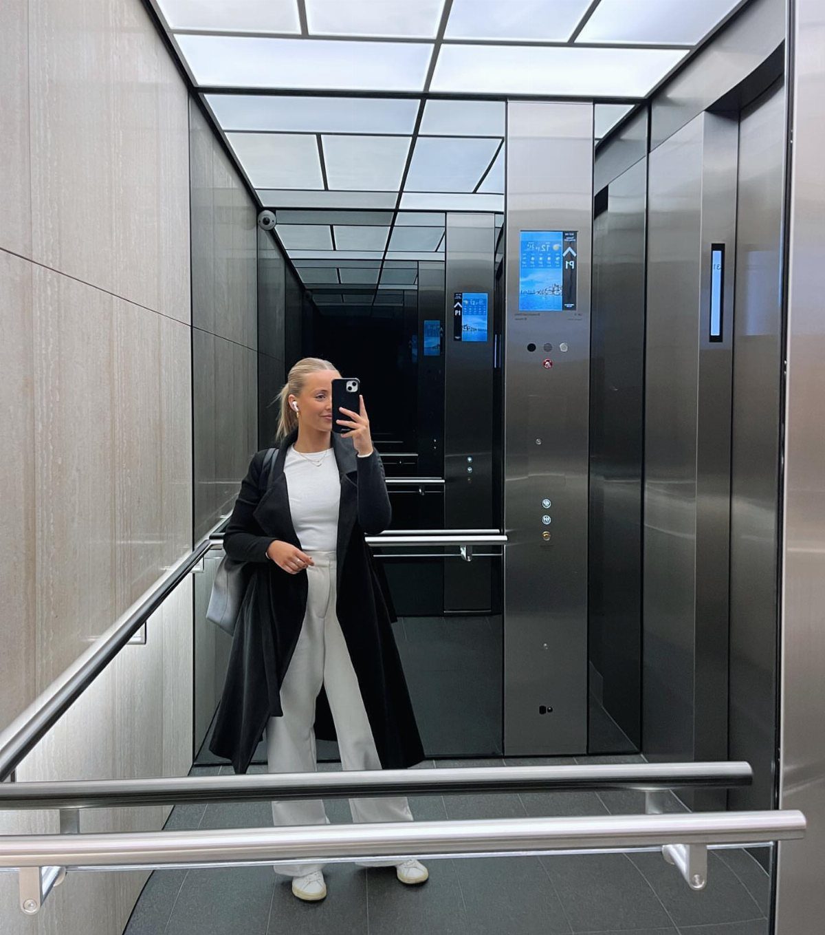 Woman in a long black overcoat and white trousers taking a mirror selfie.