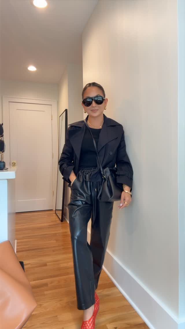 A woman wearing black faux leather joggers, a black jacket, and red heels posing in a hallway.
