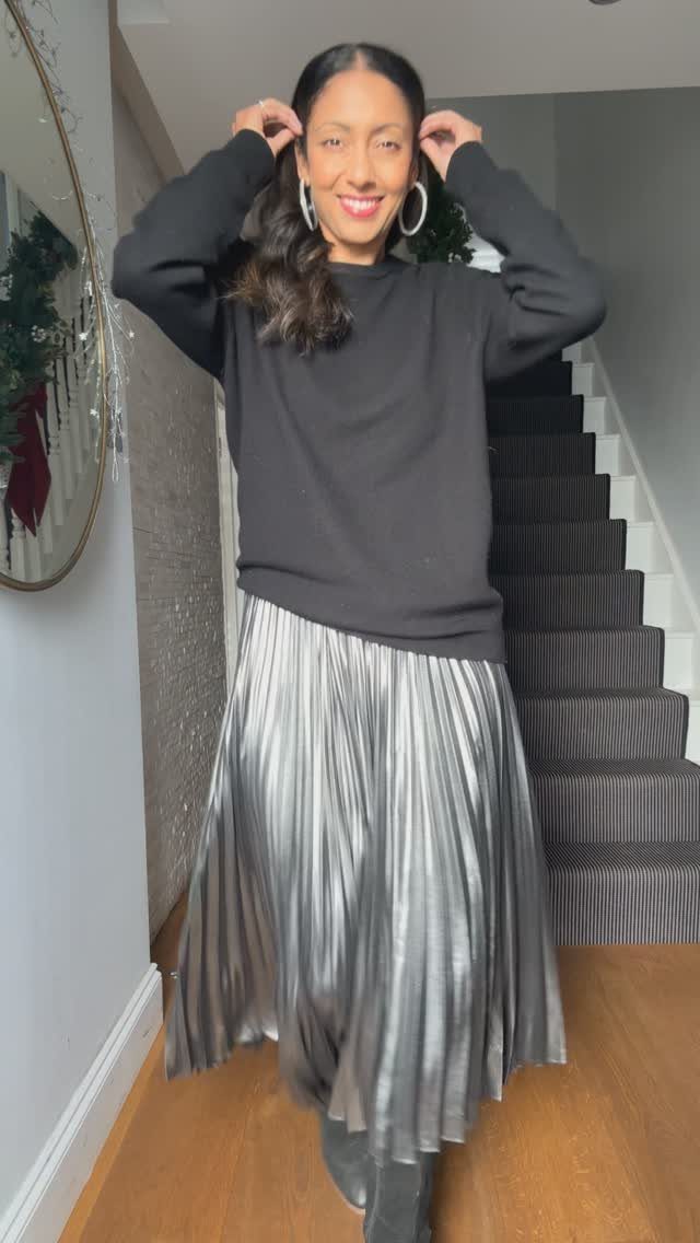 A woman wearing a black sweater and a silver metallic pleated midi skirt standing in a hallway.