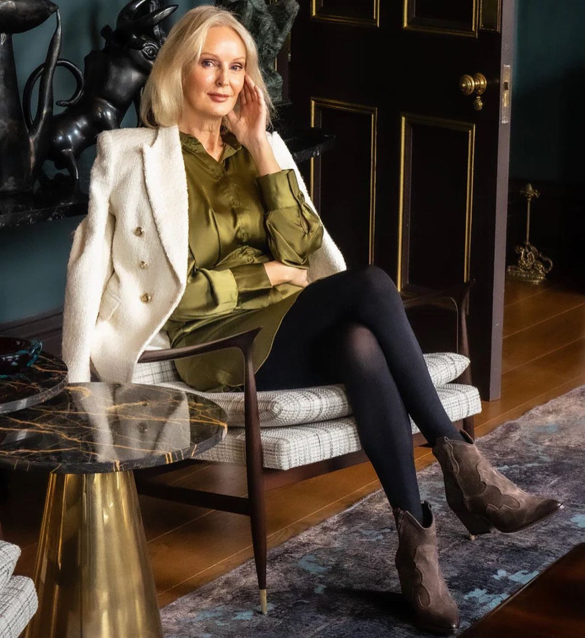 Blonde woman in olive satin blouse, white blazer, black tights, and brown suede ankle boots.