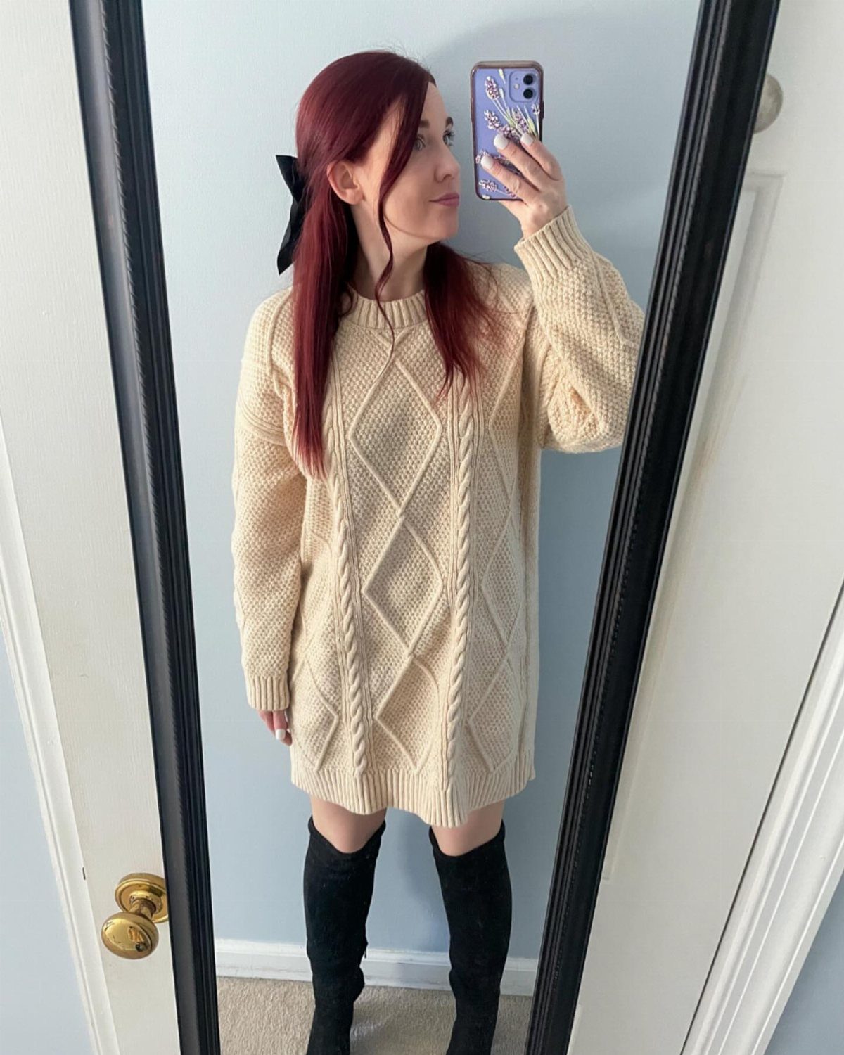 Woman in a cream cable-knit sweater dress and tall black suede boots posing in a mirror.