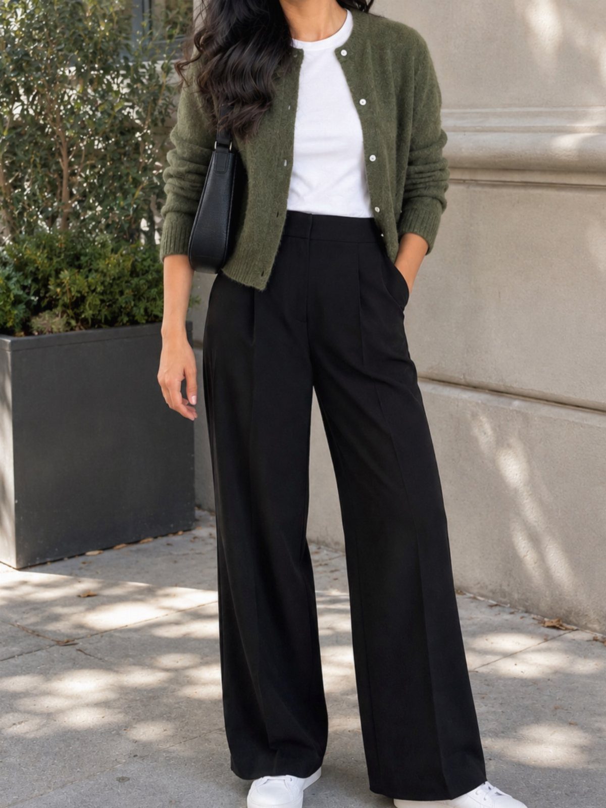 Person wearing an olive green cardigan, white t-shirt, and black wide-leg trousers standing outdoors.