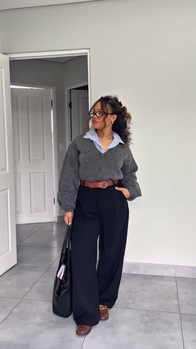 Woman wearing a grey cardigan, blue collared shirt, and black wide-leg trousers standing in a room.
