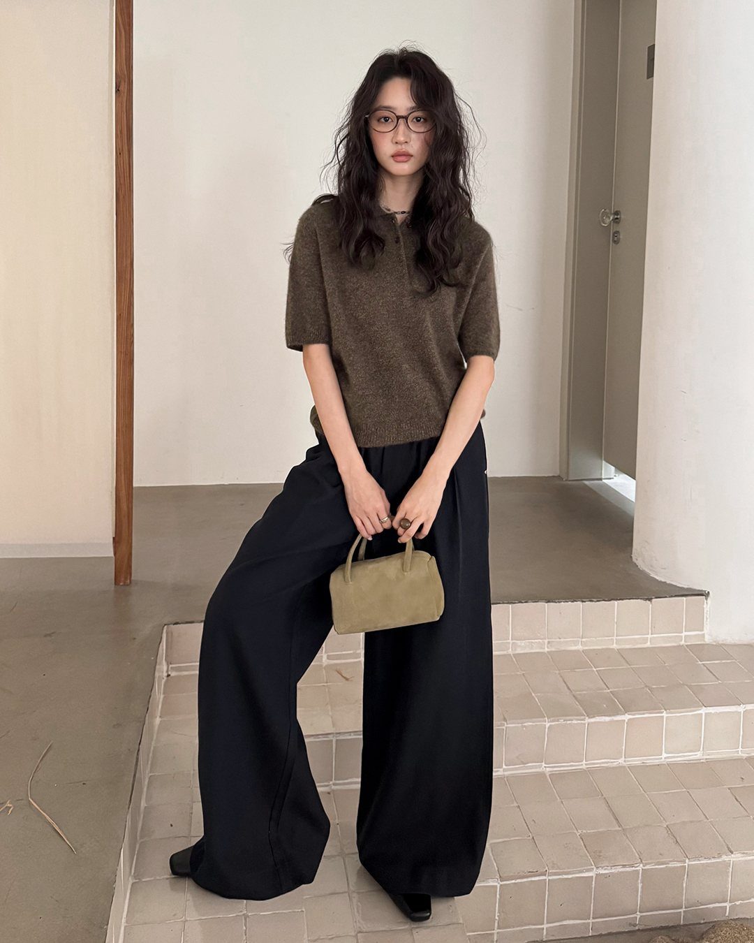 Woman wearing a brown knit polo shirt and black wide-leg trousers holding a small handbag.