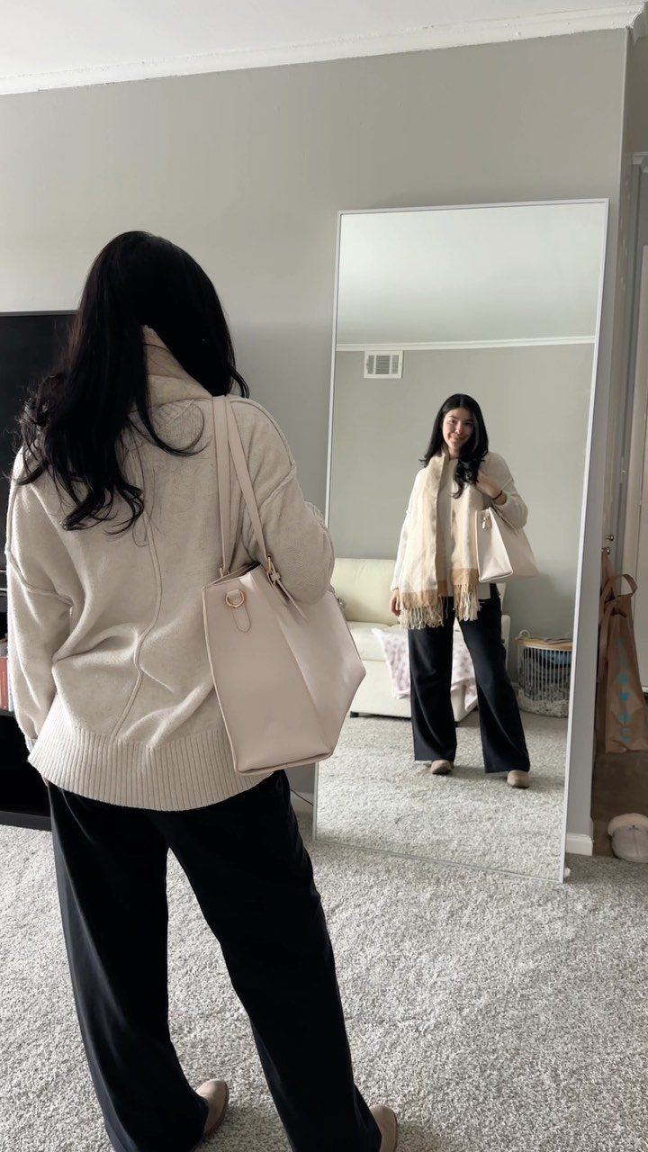 A woman in a cream sweater and black wide-leg pants posing before a mirror.