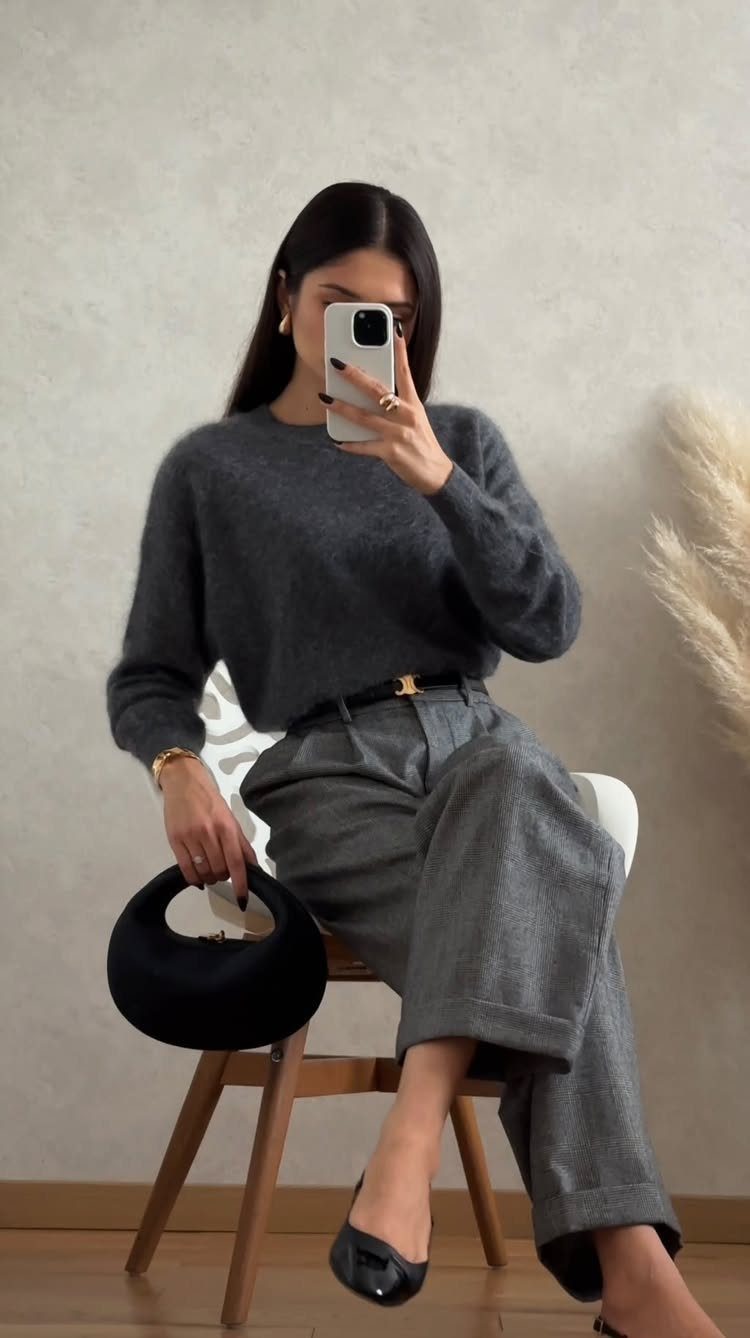 Woman in a grey sweater and wide-leg trousers sitting on a chair taking a selfie.