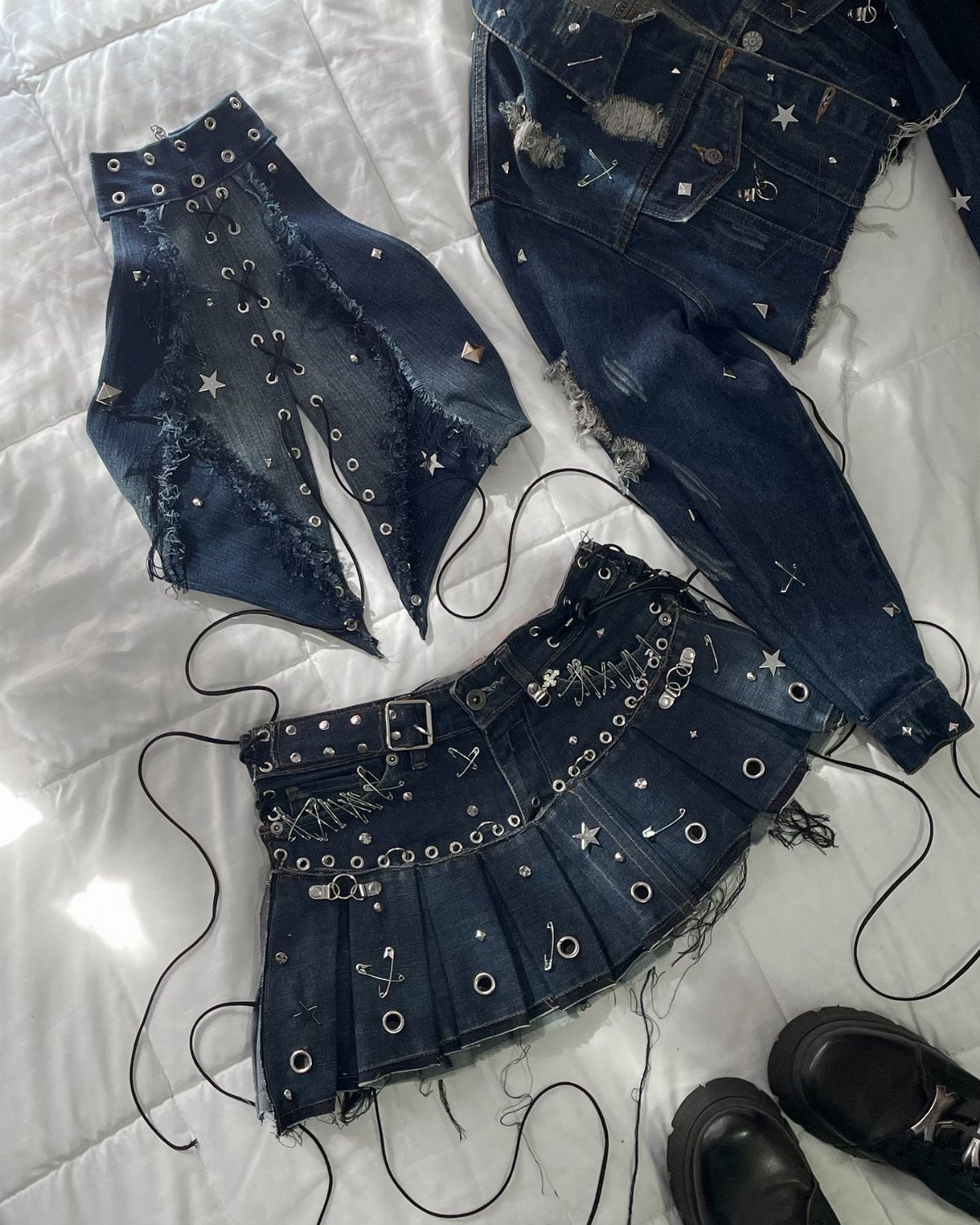 Distressed dark denim clothing items with silver grommets and star embellishments laid on white bedding.