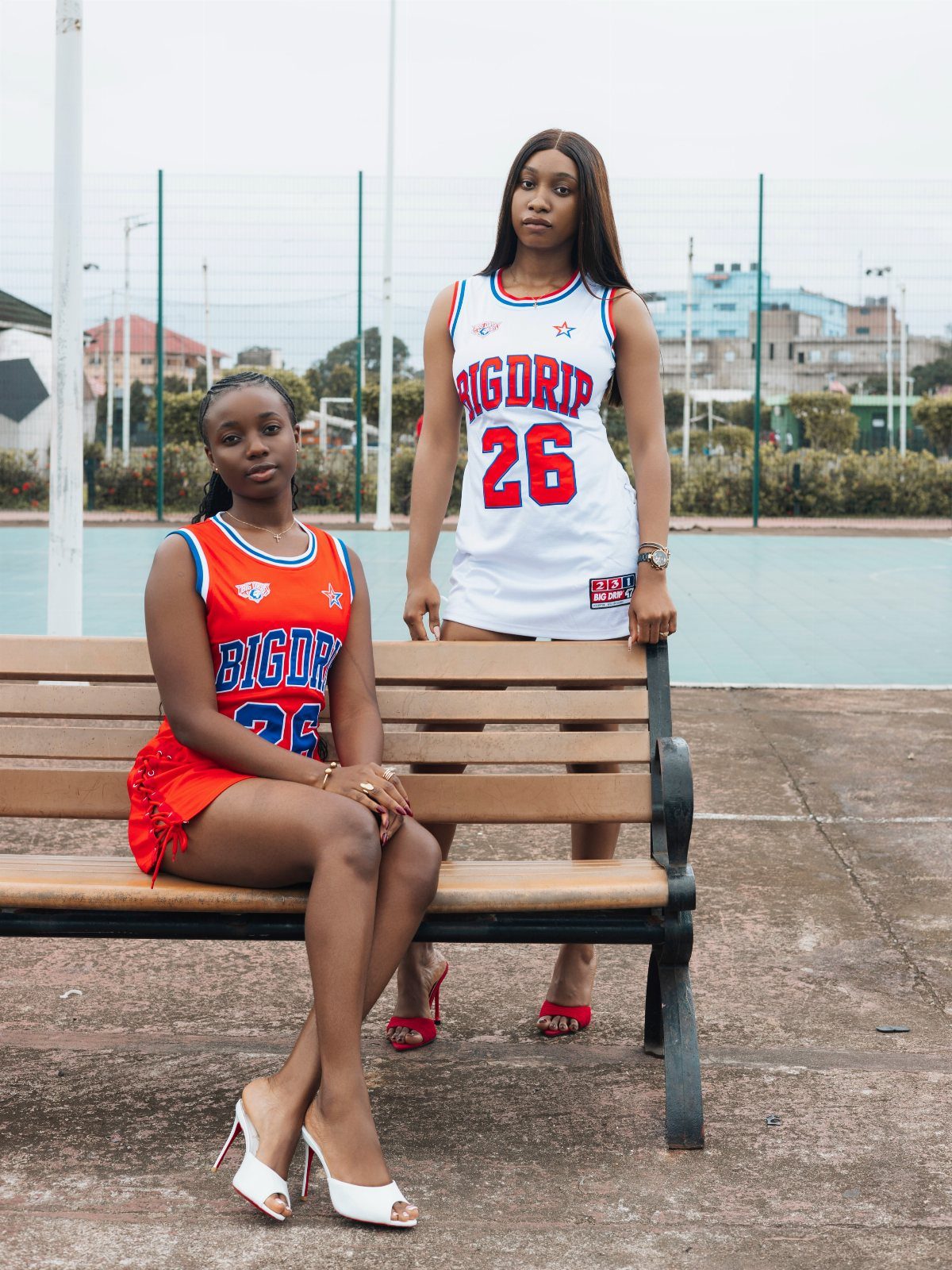 Two women posing in colorful sleeveless jersey dresses outdoors near a sports court.