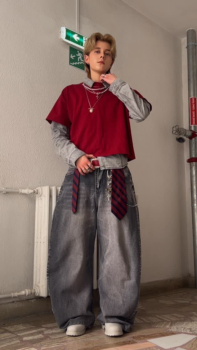 A person wearing baggy grey jeans, a red layered shirt, and multiple necklaces standing indoors.